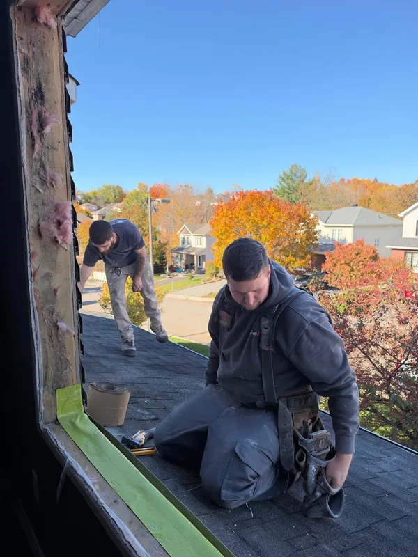 Deux installateurs près d’une ouverture de fenêtre bordée de membrane, avec des arbres aux couleurs d’automne à l’extérieur.