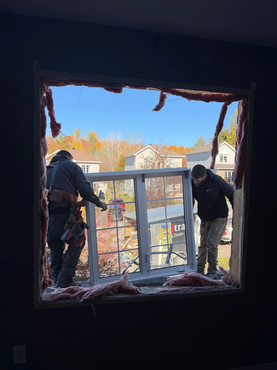 Deux installateurs manipulent une fenêtre complète dans une ouverture de mur, entre la charpente et le revêtement extérieur.