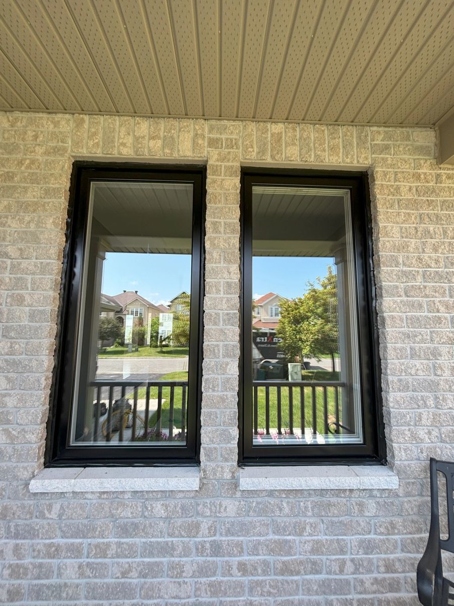 Two black-framed windows in a light brick wall, with the neighbourhood reflected in the glass.