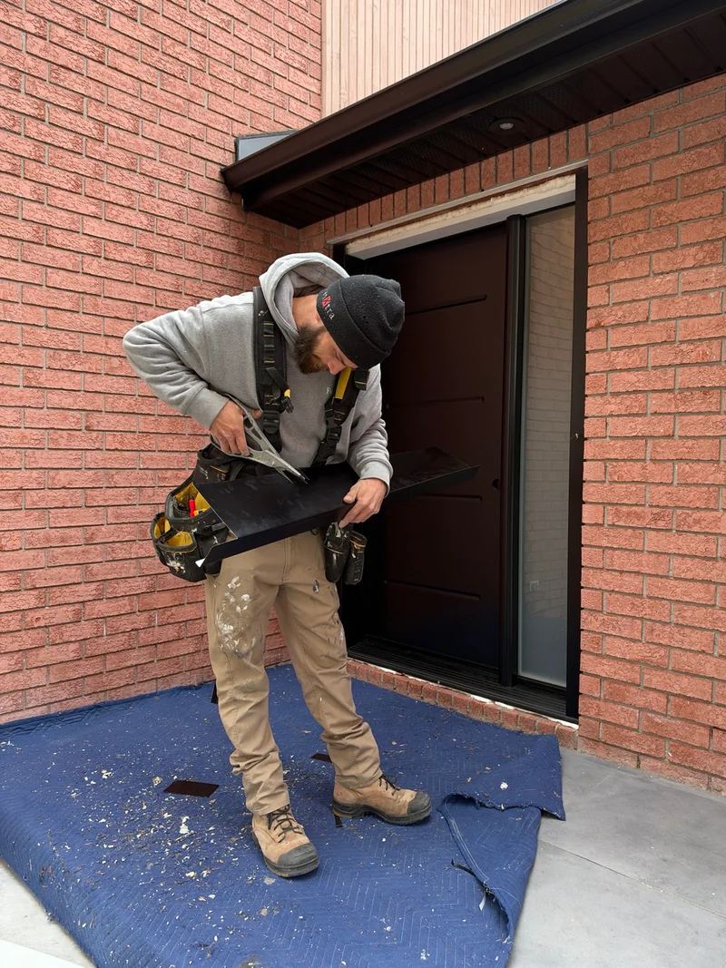 Un installateur FenXtra découpe une pièce de finition noire devant une porte, les mains et l’outil visibles.