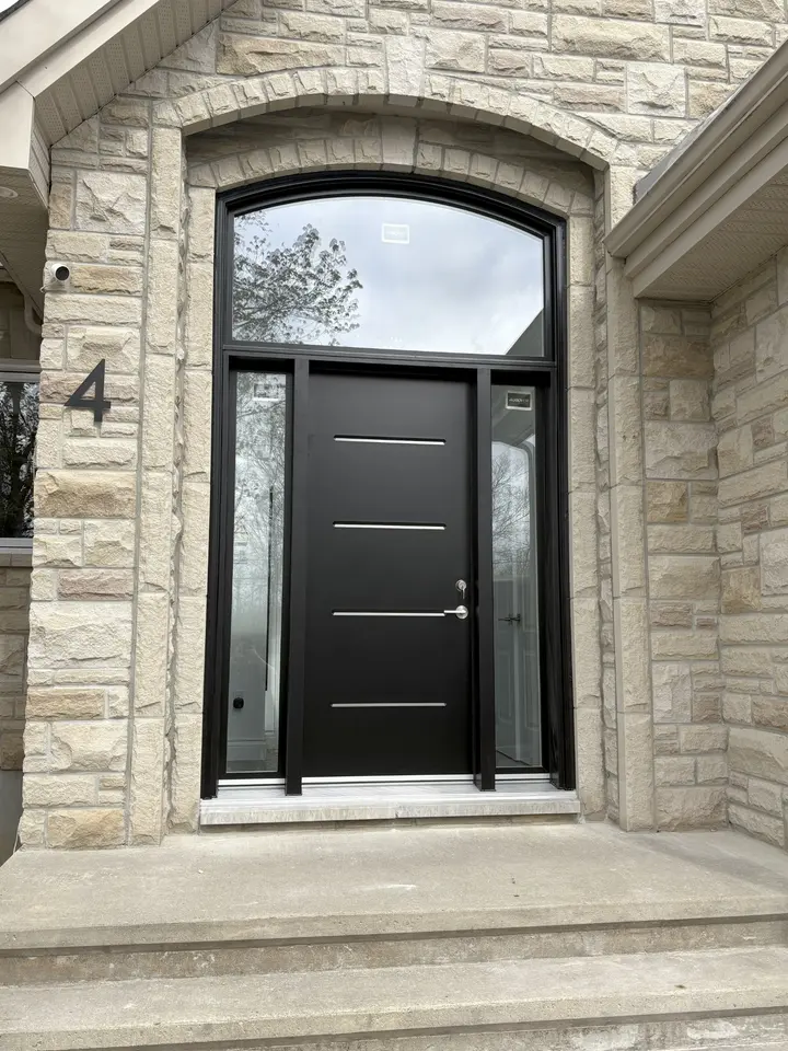 Porte d’entrée noire avec deux vitrages latéraux et une large imposte cintrée dans une façade de pierre à Val-des-Monts.